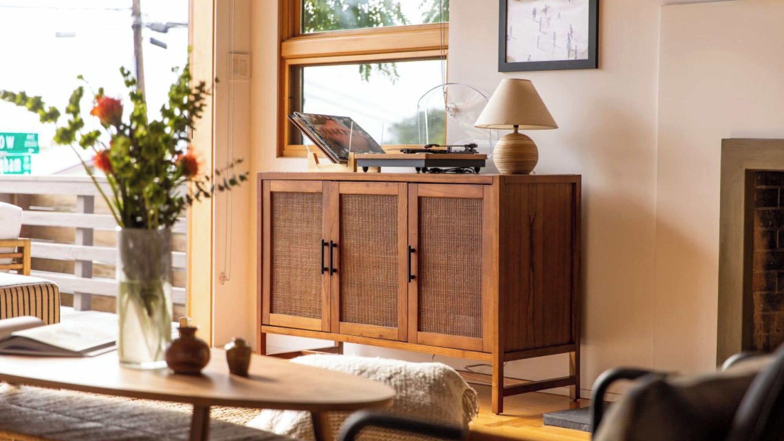 A wooden cabinet with a woven front panel, a table with a vase of flowers, and a lamp on a windowsill.