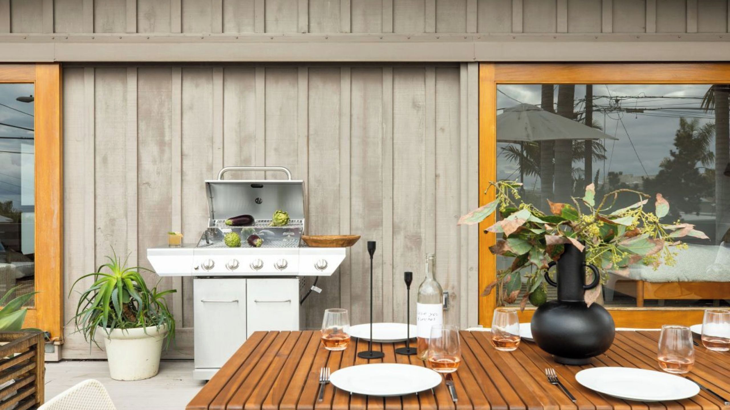 An outdoor dining area with a wooden table, white plates, and glasses, a grill, and a potted plant.