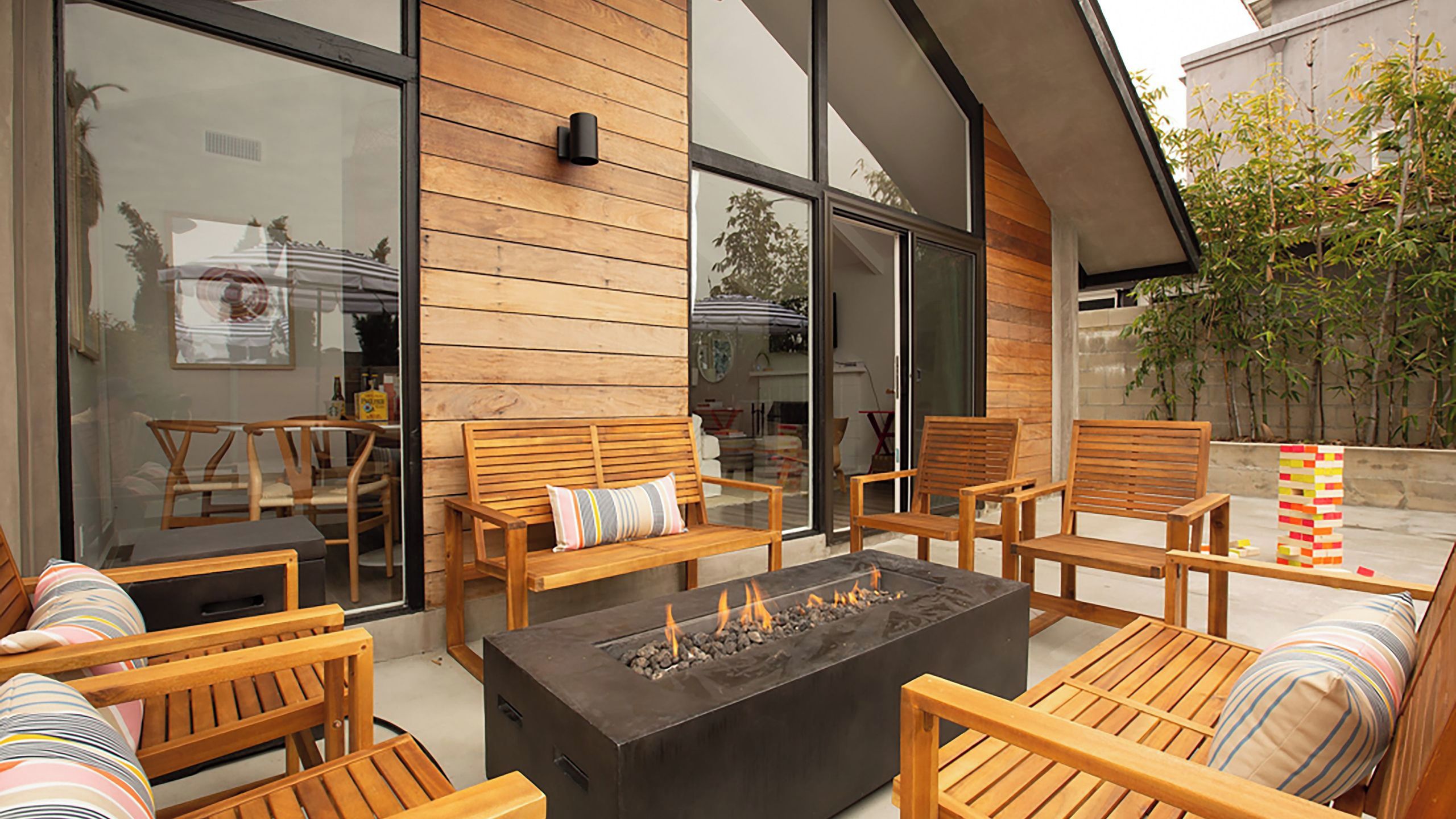 An outdoor seating area with wooden furniture, a fire pit, and a glass wall reflecting the interior.