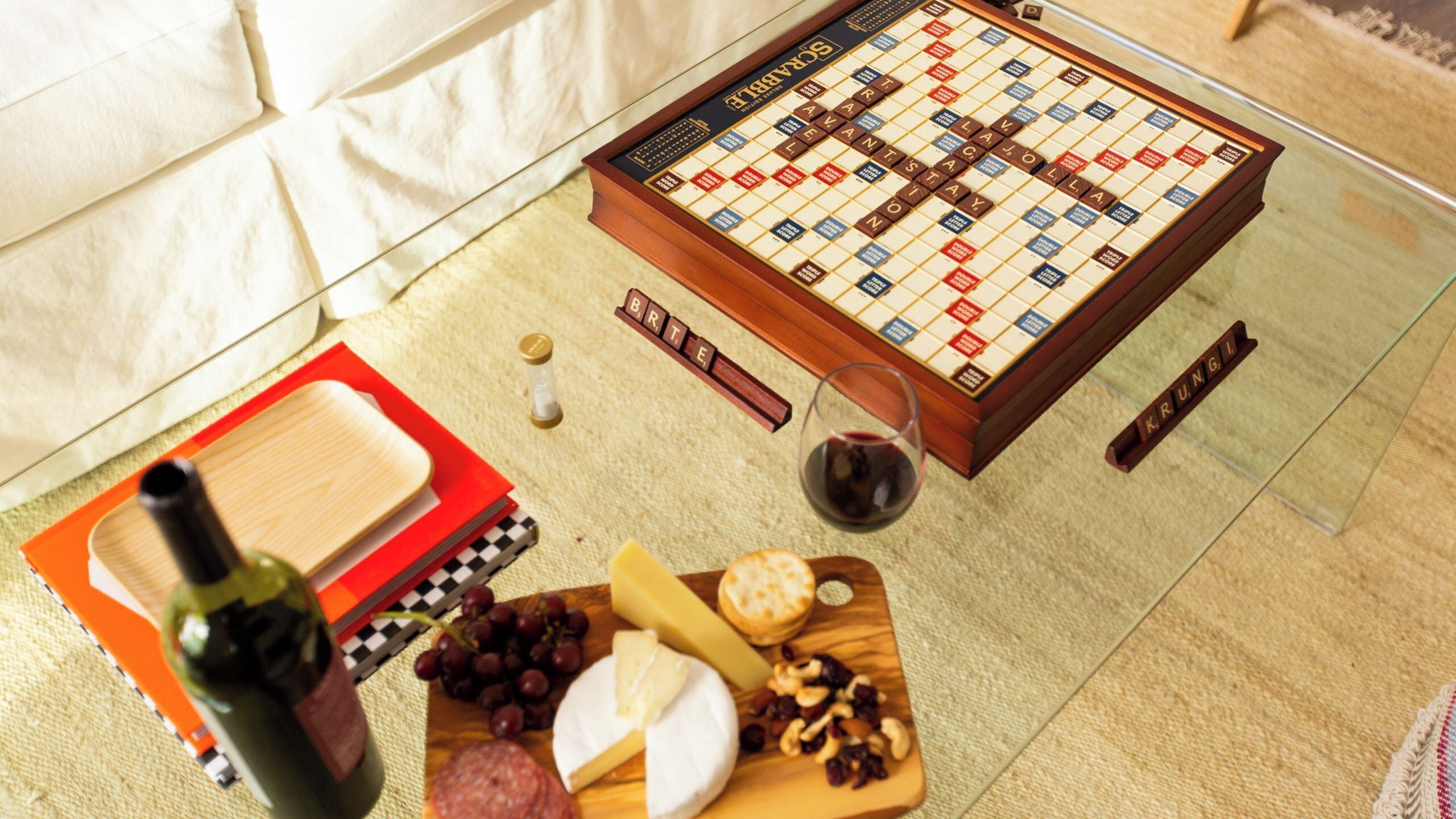 Ein Scrabble-Spiel auf einer Glastisch mit einem Brettspiel, einer Flasche, Käse, Trauben, Nüssen und einem Glas Rotwein.