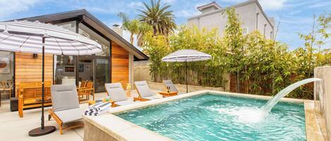 En swimmingpool med liggestole og parasoller, omgivet af grøn bevoksning og en træbygning.