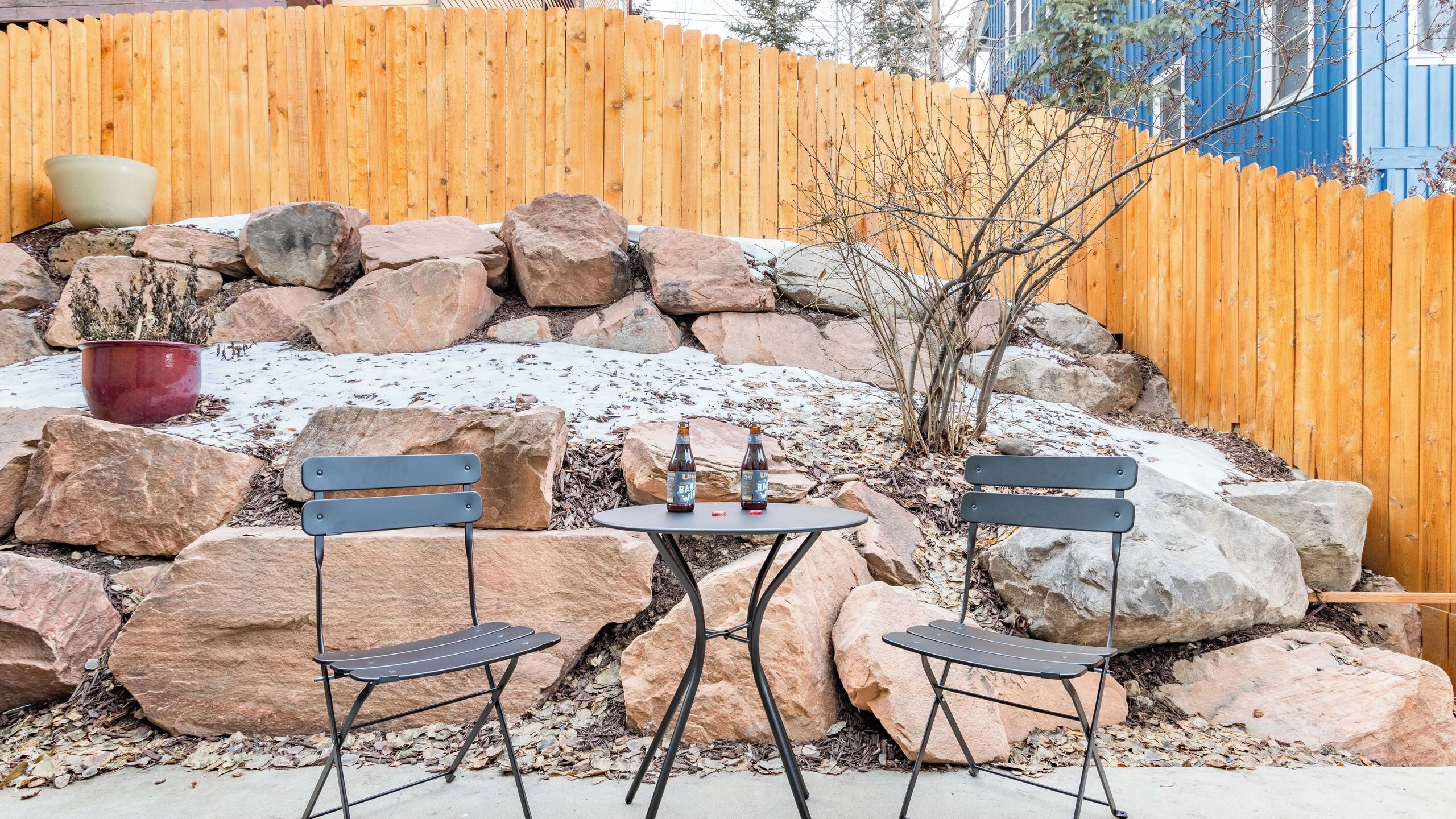 Eine Terrasse mit zwei Metallstühlen und einem kleinen runden Tisch, umgeben von großen Steinen und einem Holzzaun.