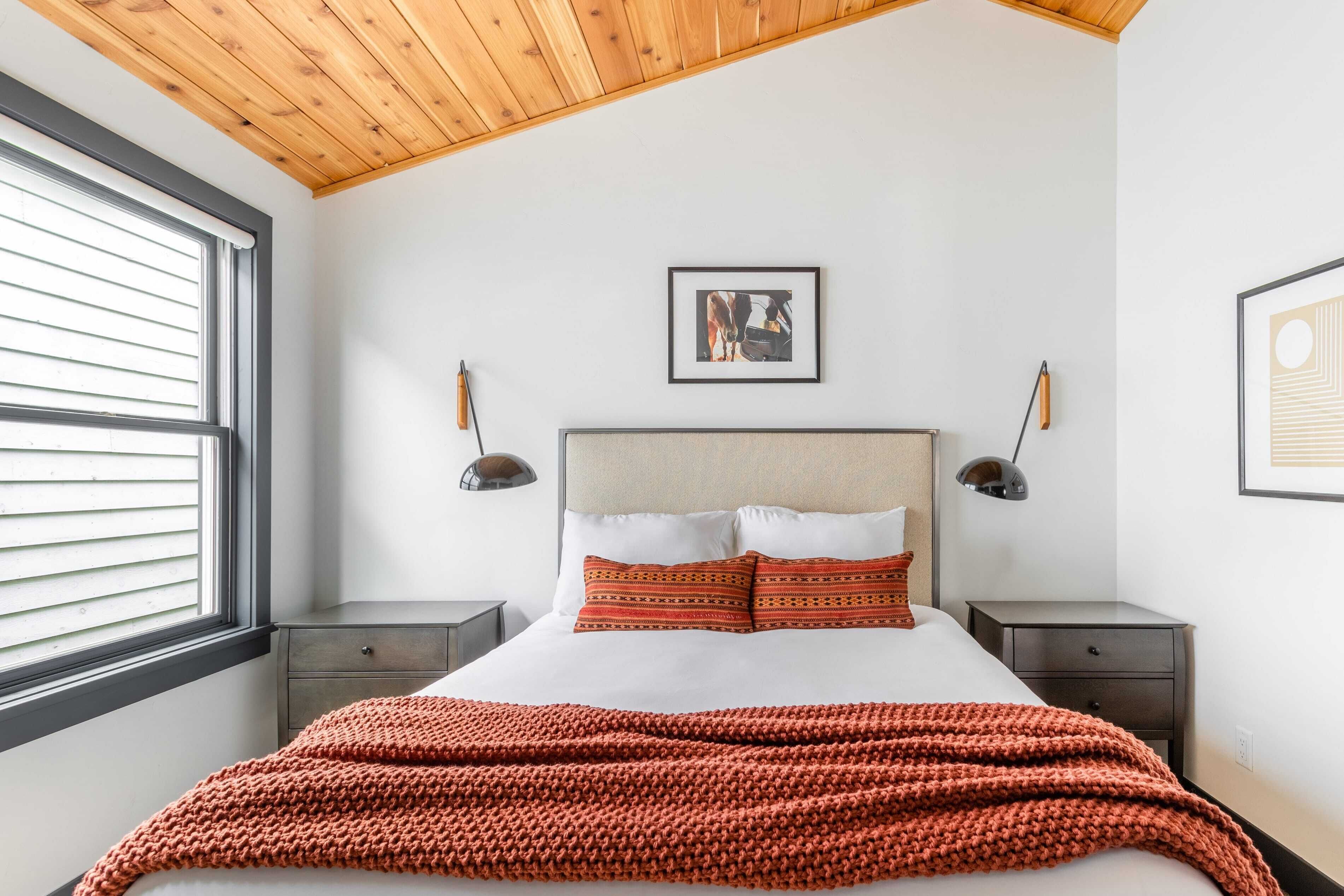 A bedroom with a wooden ceiling, a large bed with patterned pillows, two bedside tables with lamps, and a window with blinds.