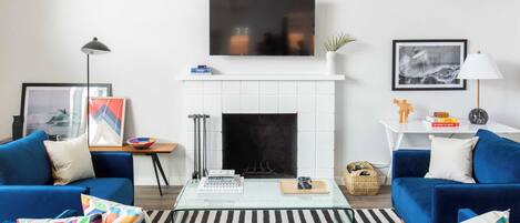 A modern living room with a fireplace, two blue sofas, a coffee table, and a flat-screen TV mounted above the fireplace.