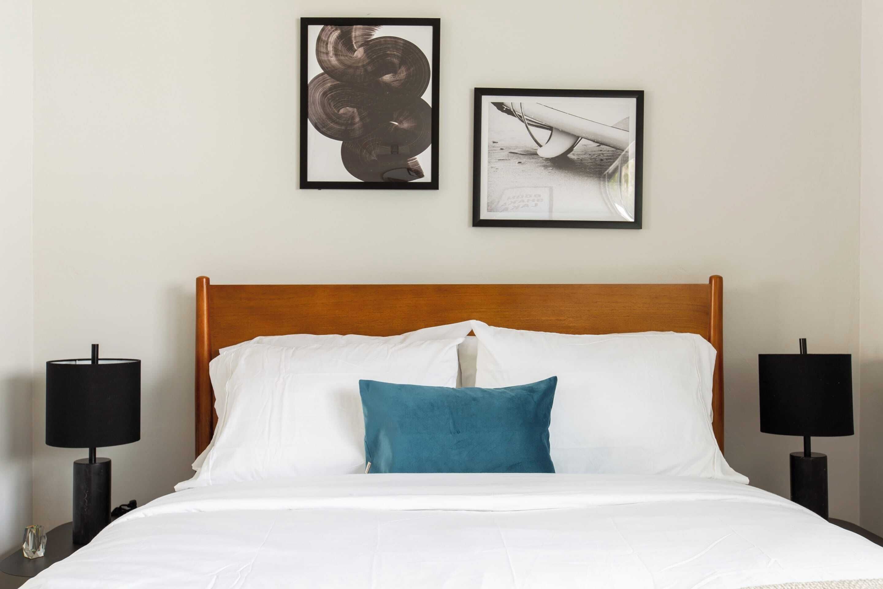 Een hotelkamer met een houten hoofdbord, witte beddengoed, een blauw-groen kussen en twee zwarte lampen aan weerszijden van het bed.