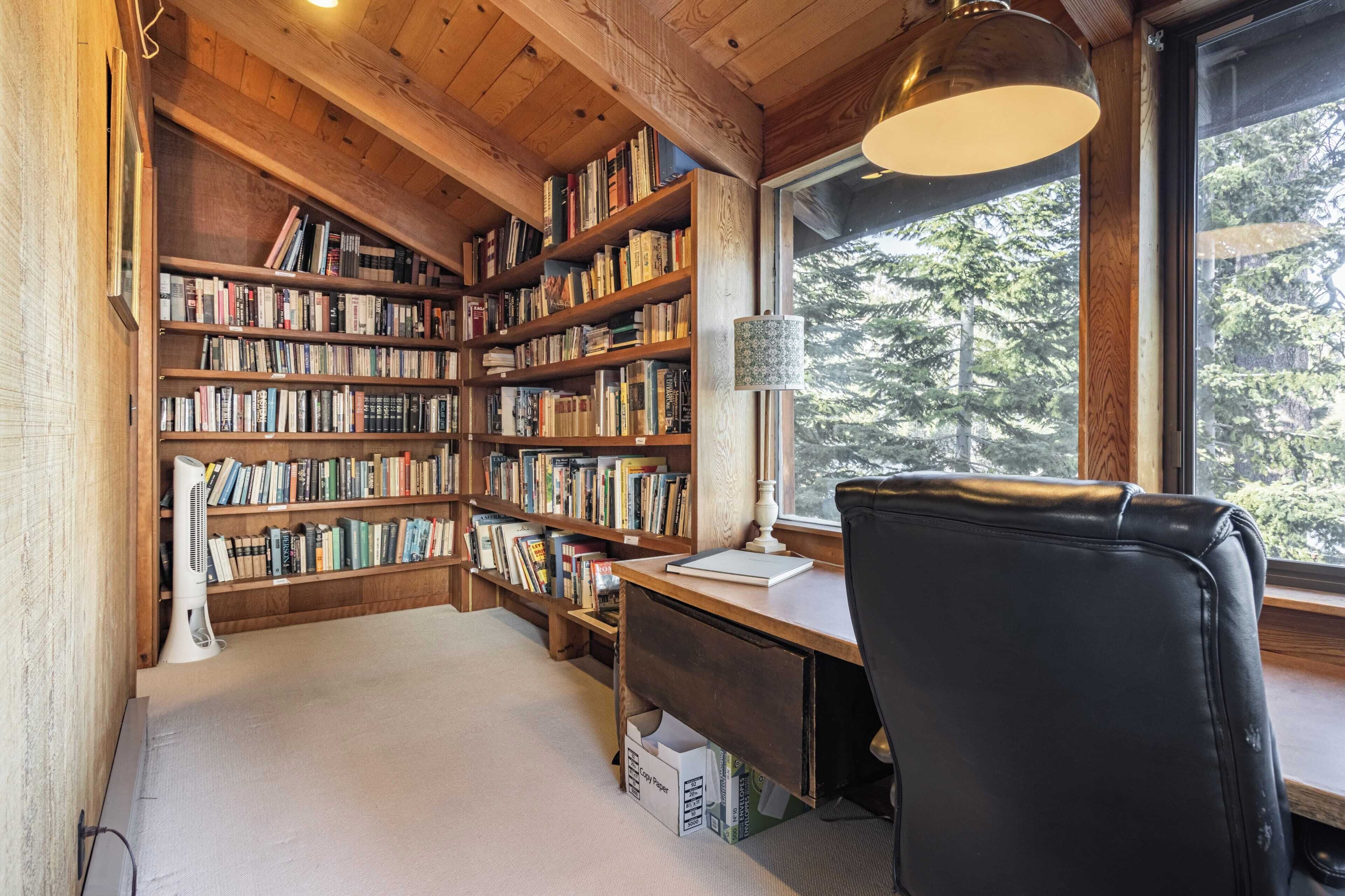 A cozy reading nook with a wooden bookshelf filled with books, a desk with a lamp, and a window offering a view of trees.