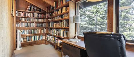 A cozy reading nook with a wooden bookshelf filled with books, a desk with a lamp, and a window offering a view of trees.
