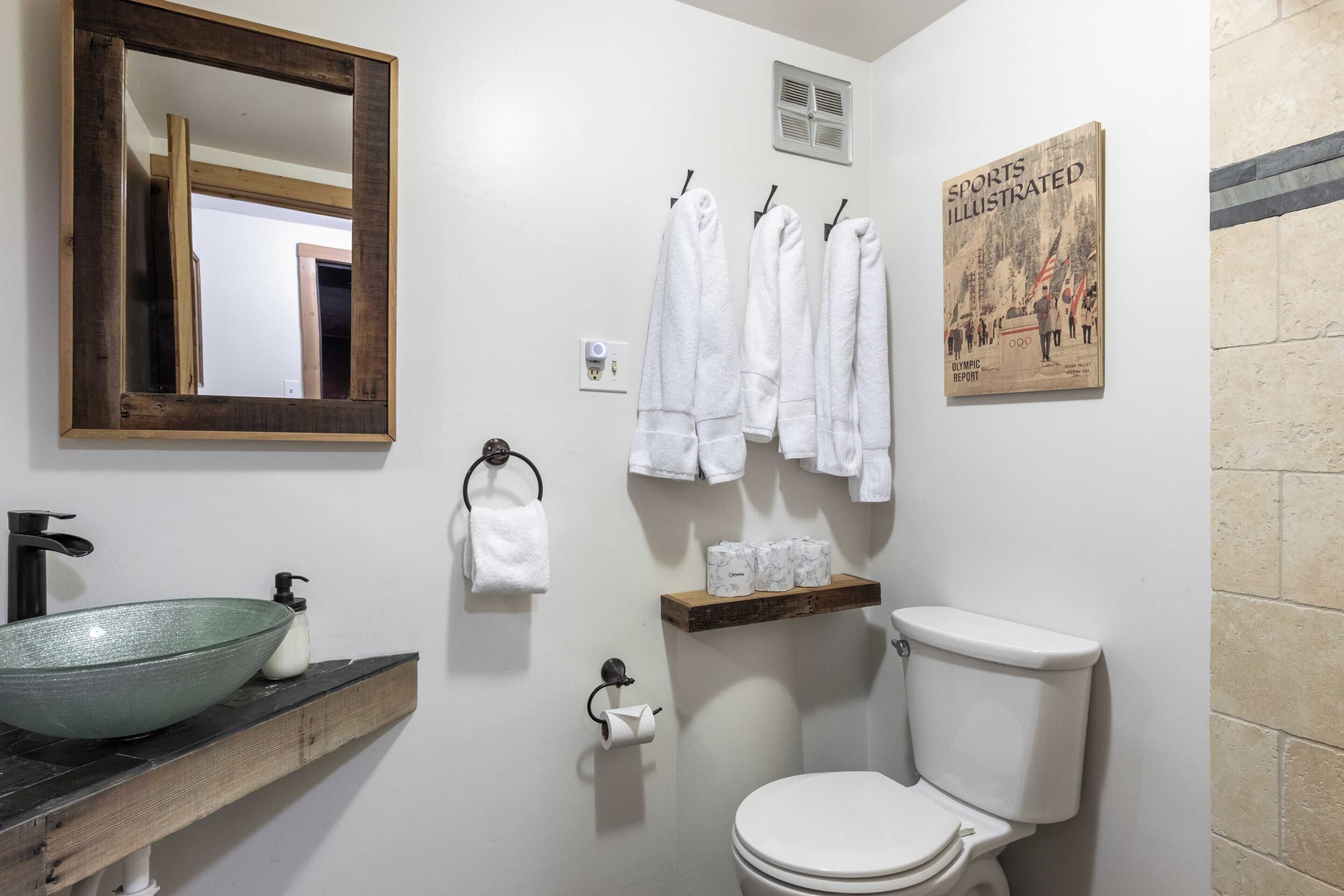 A bathroom with a large mirror, a glass bowl sink, a toilet, and a shelf with towels and toilet paper.
