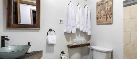 A bathroom with a large mirror, a glass bowl sink, a toilet, and a shelf with towels and toilet paper.