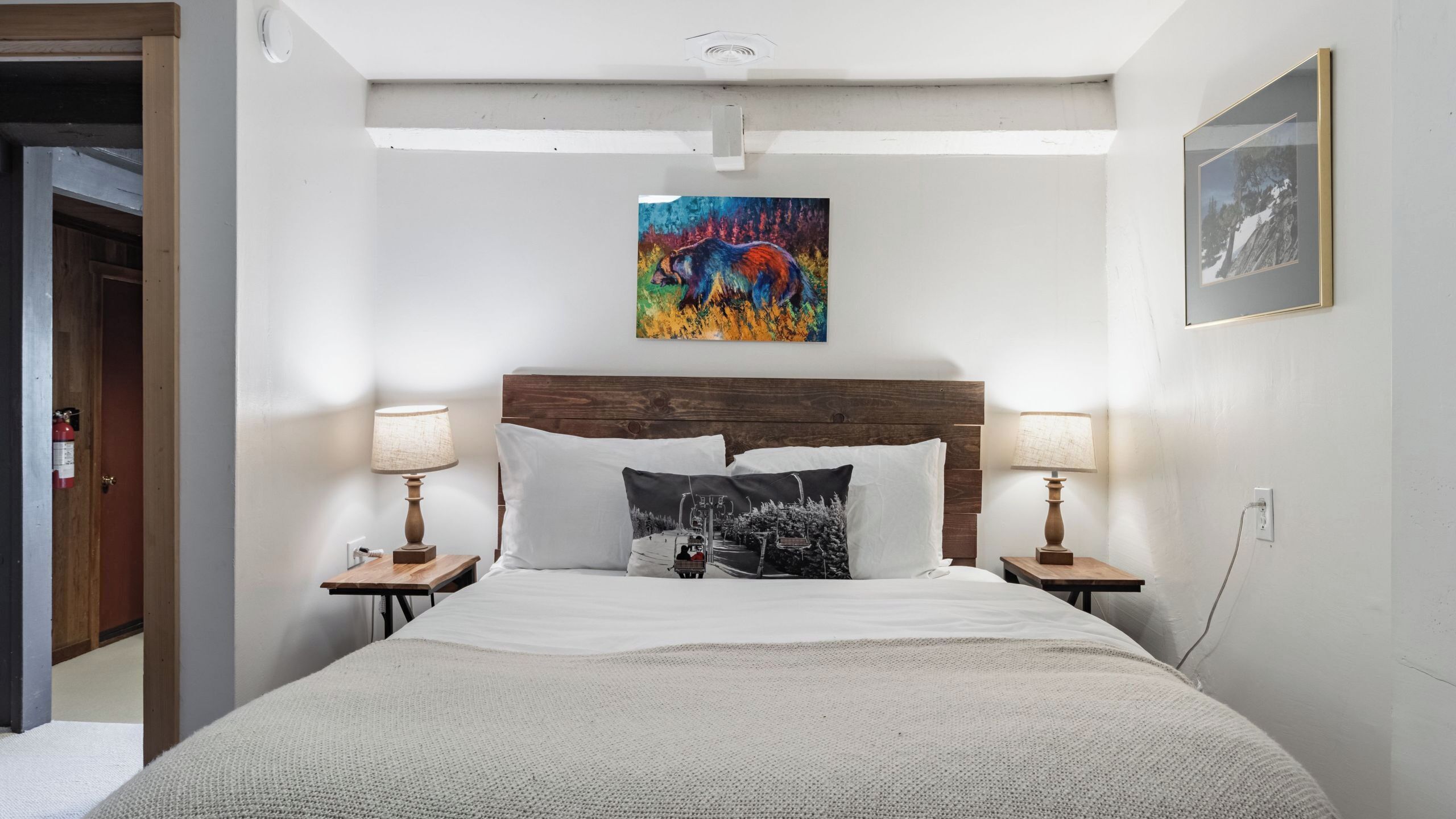 Habitación de hotel con cabecera de madera, cama con ropa de cama blanca, dos lámparas de noche, un cuadro abstracto colorido sobre la cama y una fotografía enmarcada en la pared.