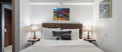 Habitación de hotel con cabecera de madera, cama con ropa de cama blanca, dos lámparas de noche, un cuadro abstracto colorido sobre la cama y una fotografía enmarcada en la pared.