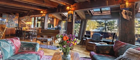 L’intérieur chaleureux d’un chalet avec des poutres en bois, un coin repas et une grande fenêtre donnant sur la forêt.