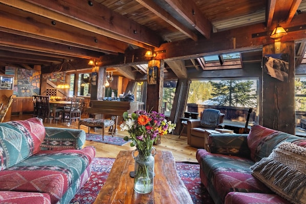 A cozy cabin interior with wooden beams, a dining area, and a large window with a view of trees.