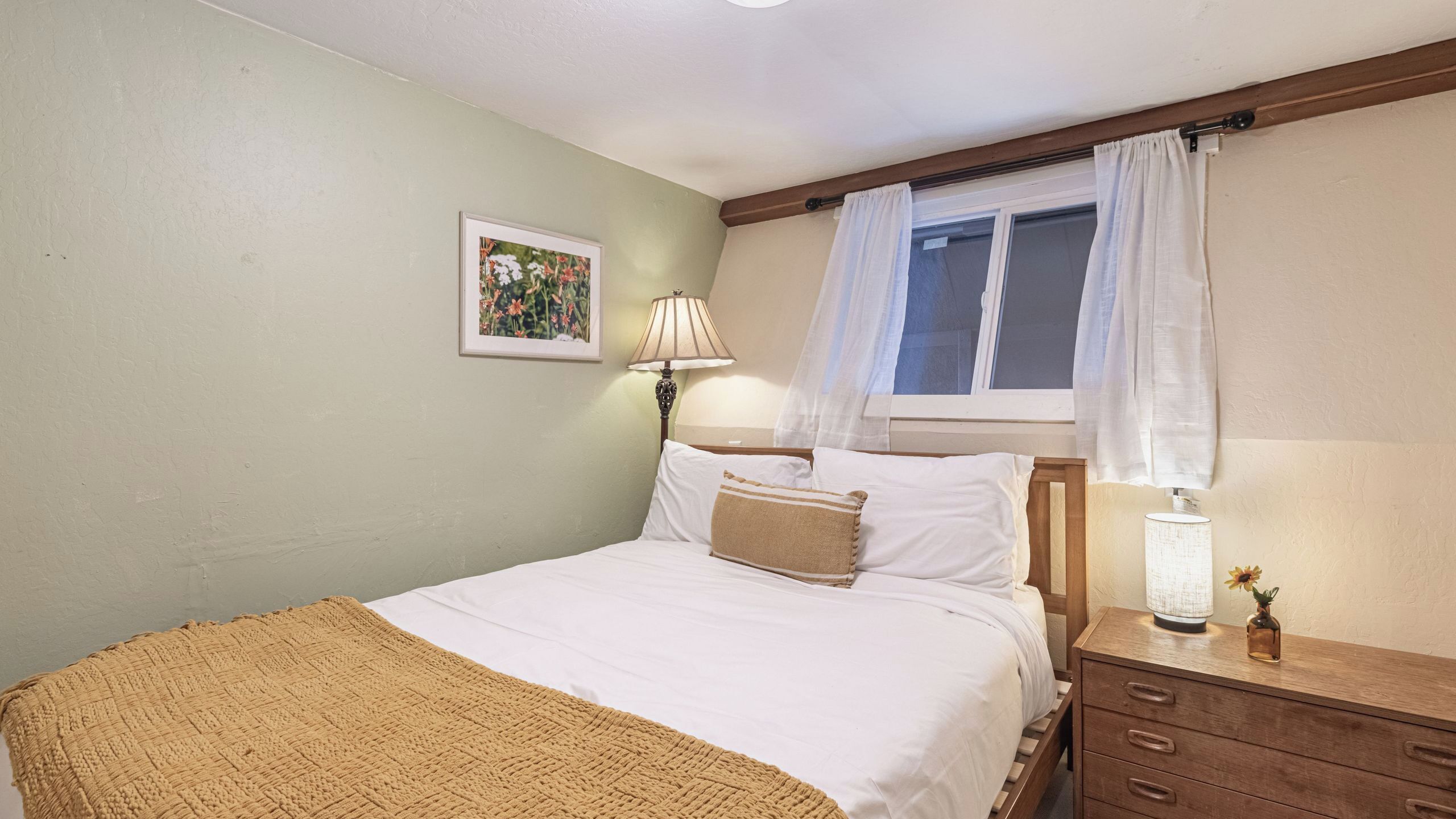 A bedroom with a bed, a bedside table with a lamp, a window with curtains, a wall-mounted picture, and a wooden headboard.