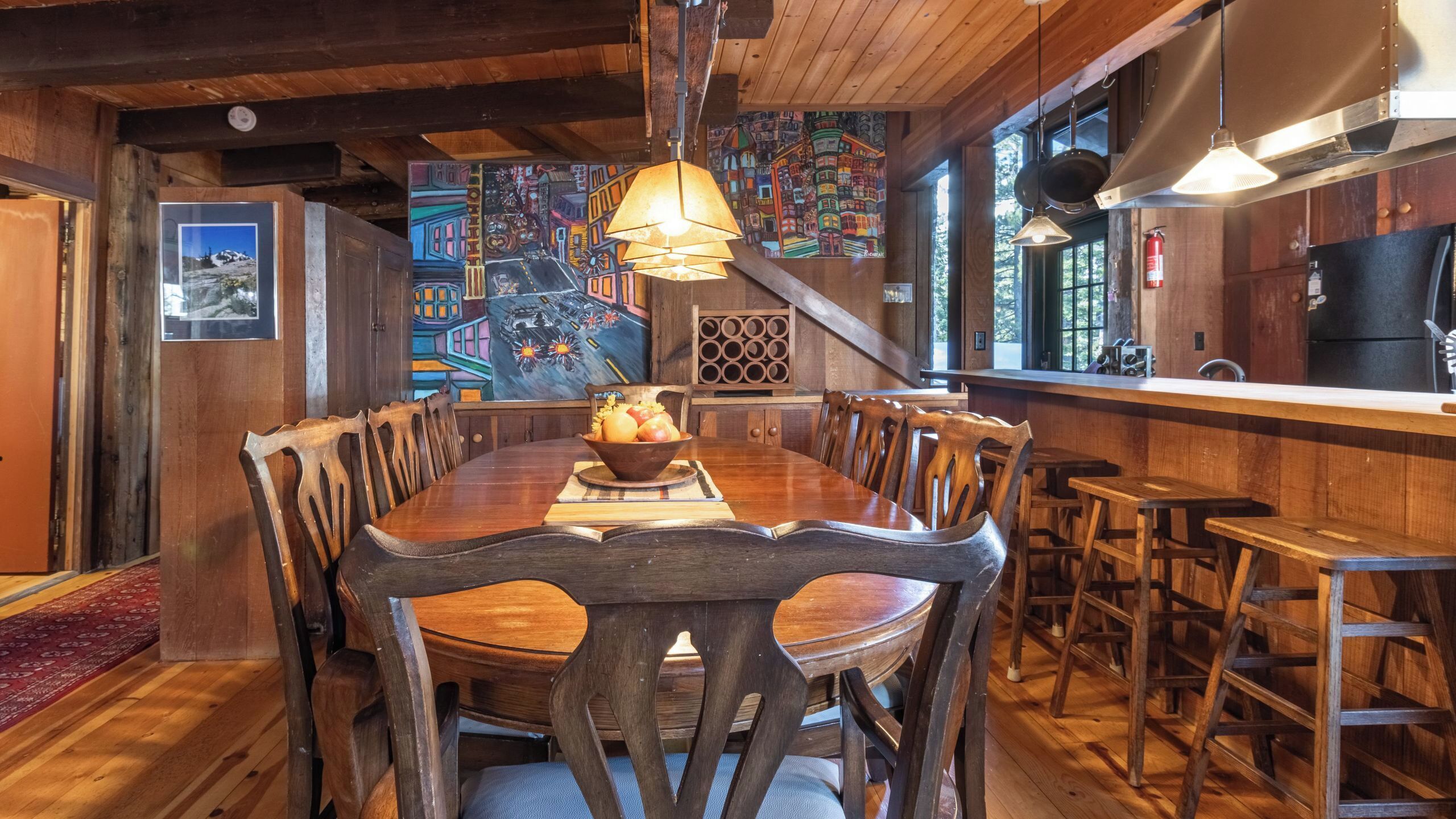 A wooden dining table with chairs, a bowl of fruit, and a mural on the wall.