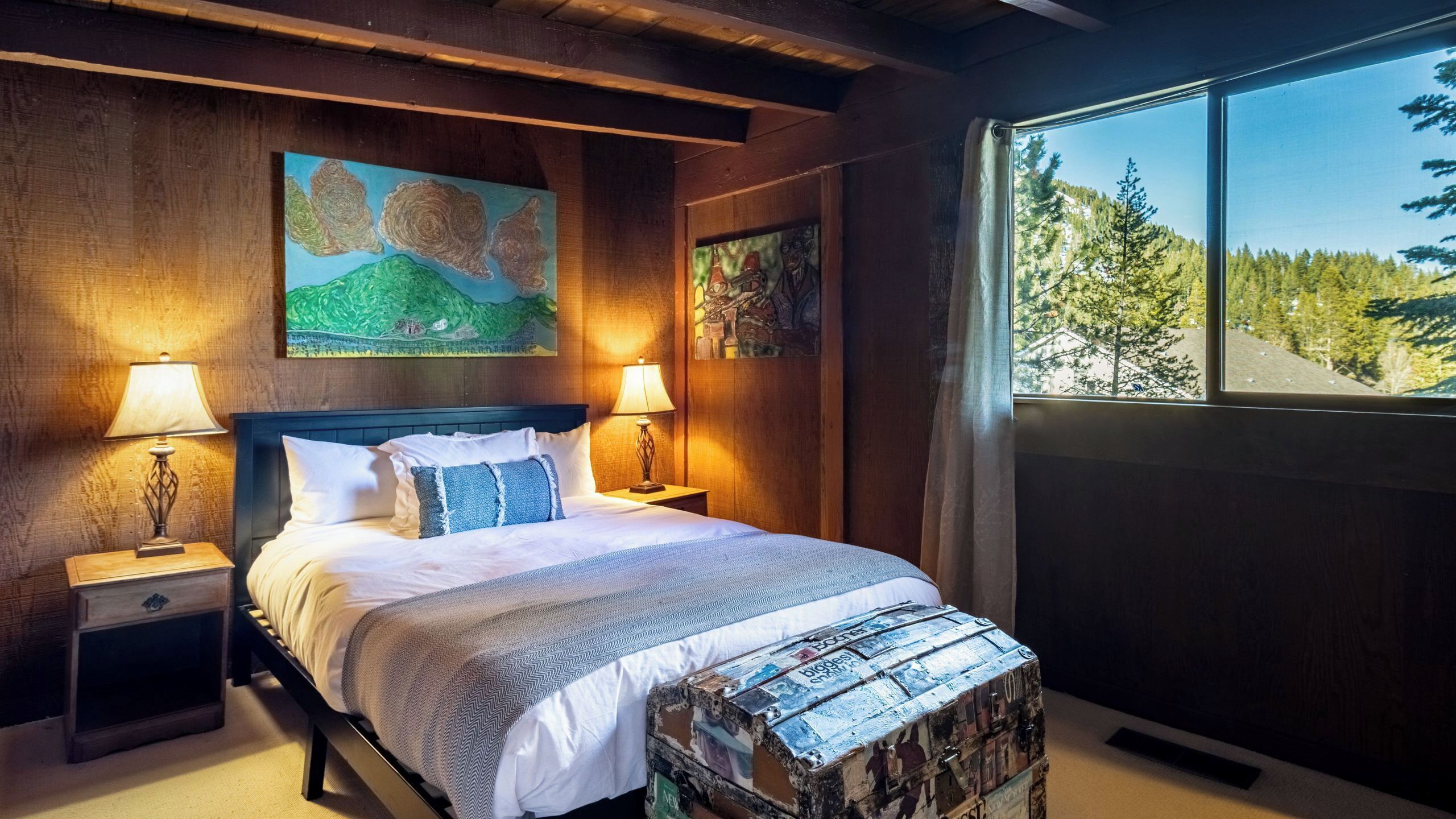 A bedroom with a bed, two bedside tables with lamps, a large window with a forest view, and a painting on the wall.
