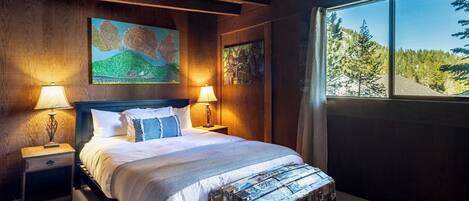 A bedroom with a bed, two bedside tables with lamps, a large window with a forest view, and a painting on the wall.