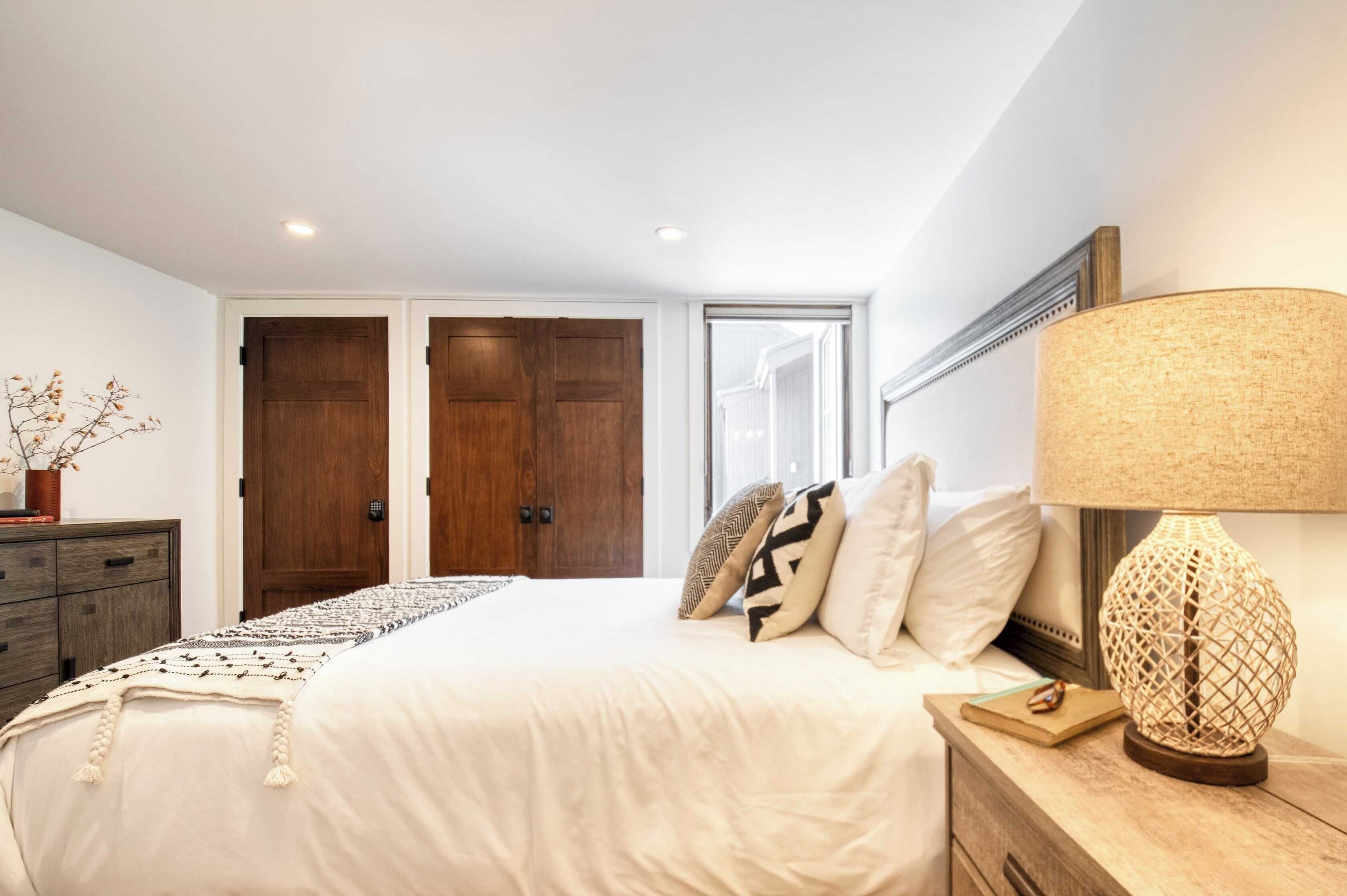 A bedroom with a large bed, a nightstand with a lamp, a dresser, and a door leading to another room.