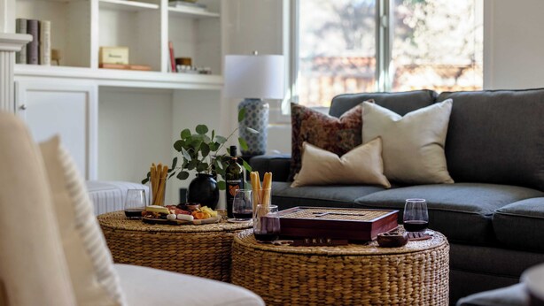 A cozy living room with a grey sofa, wicker ottomans, and a bookshelf filled with books and decorative items. - Cabernet By Avantstay Tranquil Sonoma Valley Oasis w/ Pool & Fire Pit (Santa Rosa)
