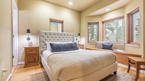 A bedroom with a large bed, wooden nightstands, a desk, and two windows with views of trees. - River Rock by Avantstay 8 Bedroom Ski Estate w/ Hot Tub & Movie Theatre 10 Min Walk to Mtn (Breckenridge)