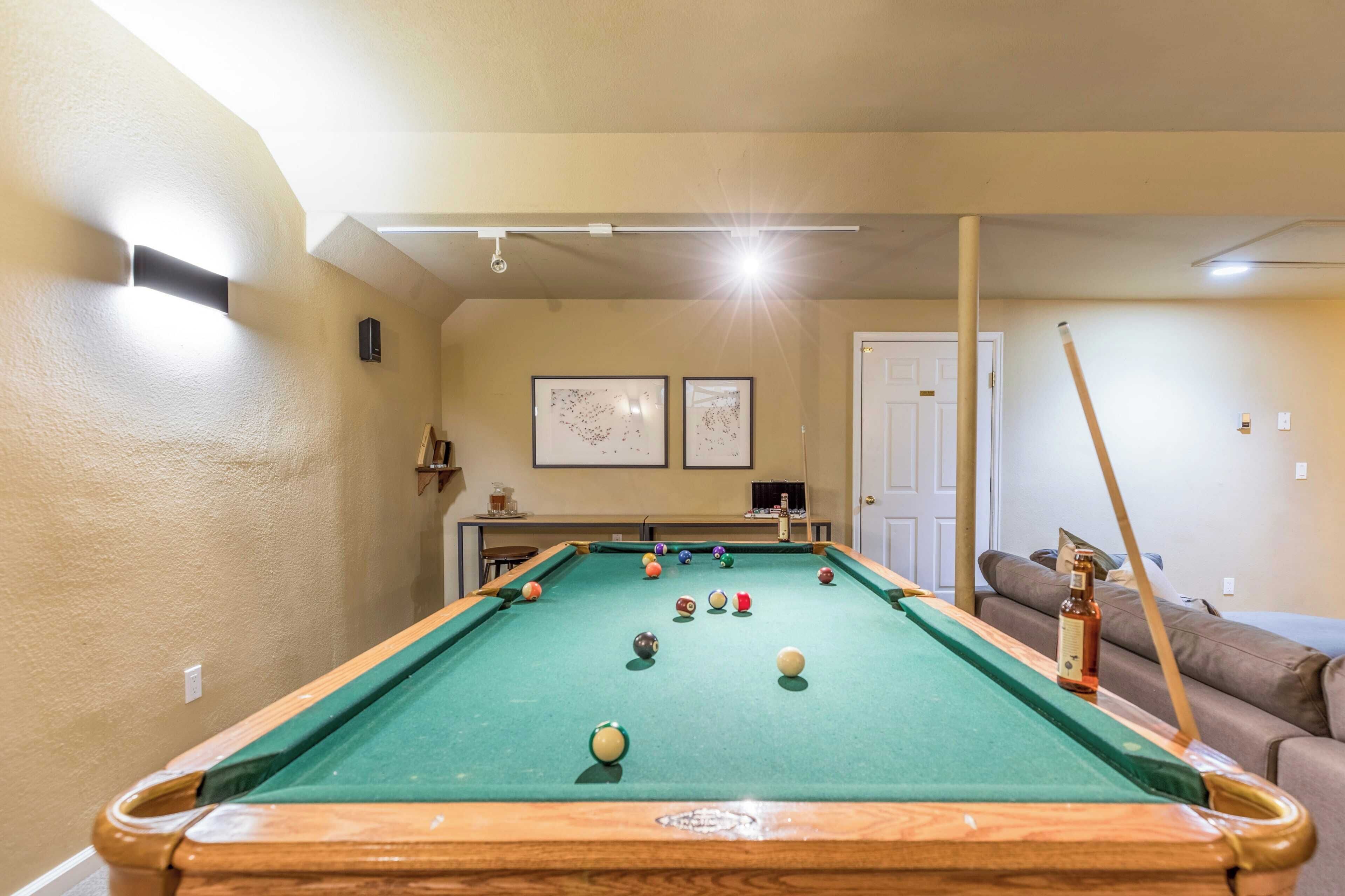 Une table de billard avec des billes, une queue de billard et une bouteille de spiritueux, dans une pièce aux murs de couleur beige et éclairée par un luminaire au plafond.