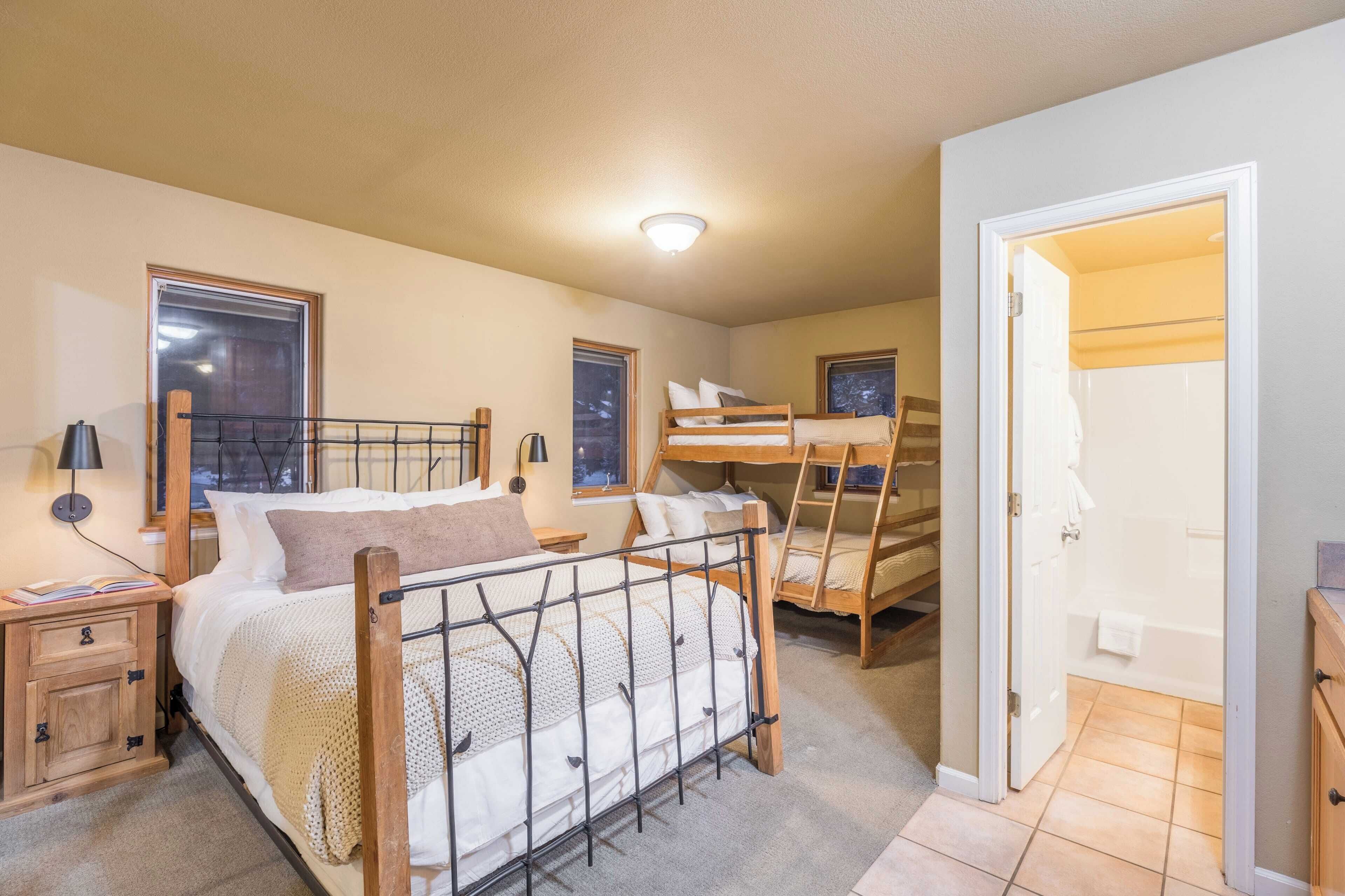 A bedroom with a bunk bed, a single bed, a nightstand with a lamp, and a door leading to a bathroom.