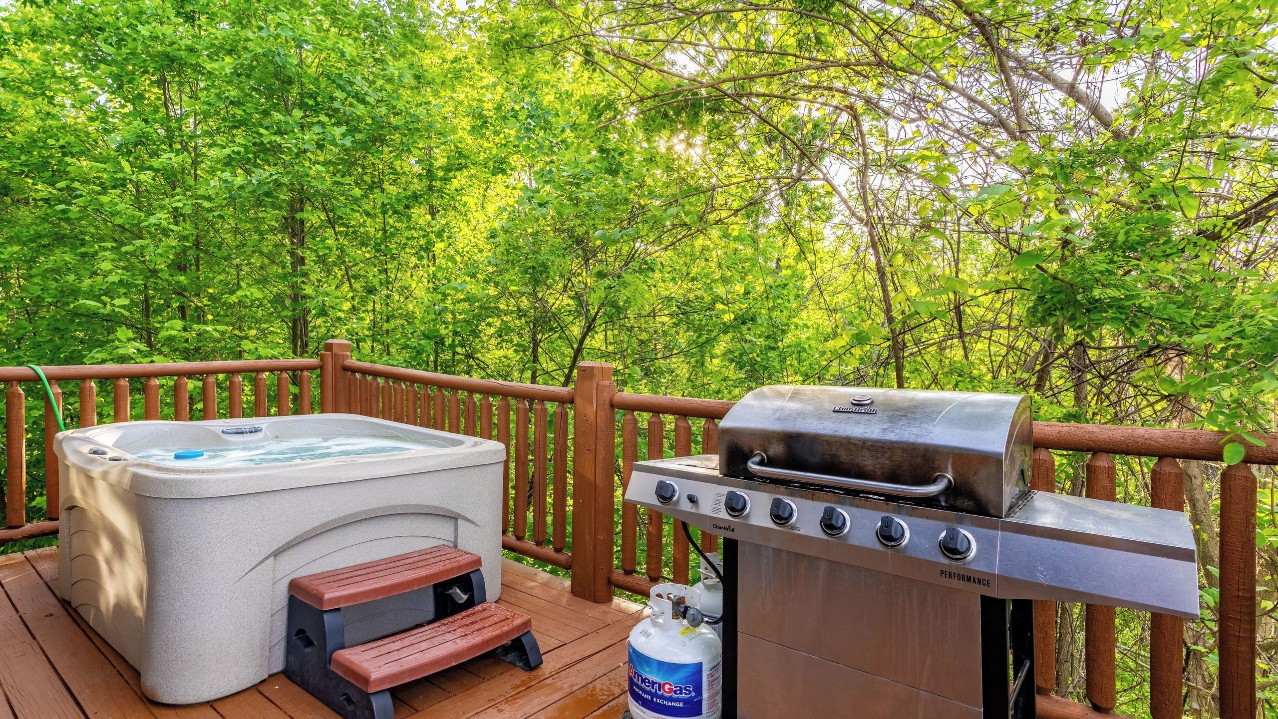 Un jacuzzi y una parrilla en una terraza exterior con barandilla de madera y abundante vegetación de fondo.