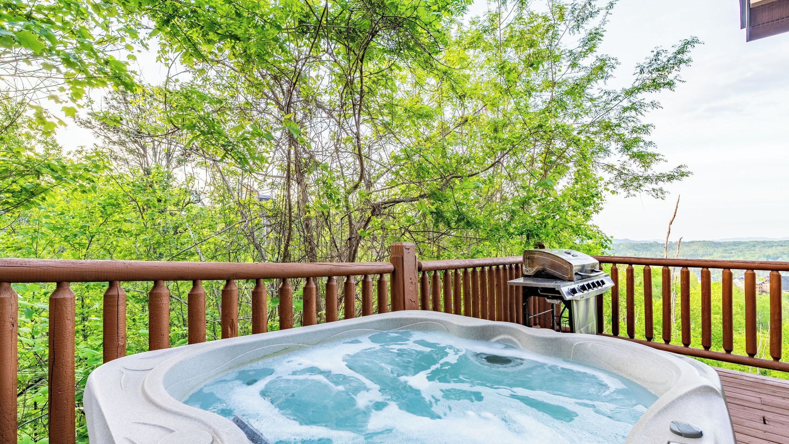 Ein Whirlpool auf einer Holzterrasse mit Blick auf Bäume und einem Grill.