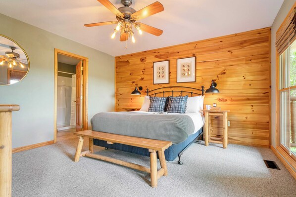 A bedroom with a wooden wall, a bed with a blue and white bedspread, a wooden bench, a bedside table with a lamp, and a ceiling fan.