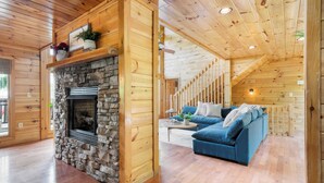 A log cabin interior with a stone fireplace, wooden walls, a blue sofa, and a staircase leading to an upper level.