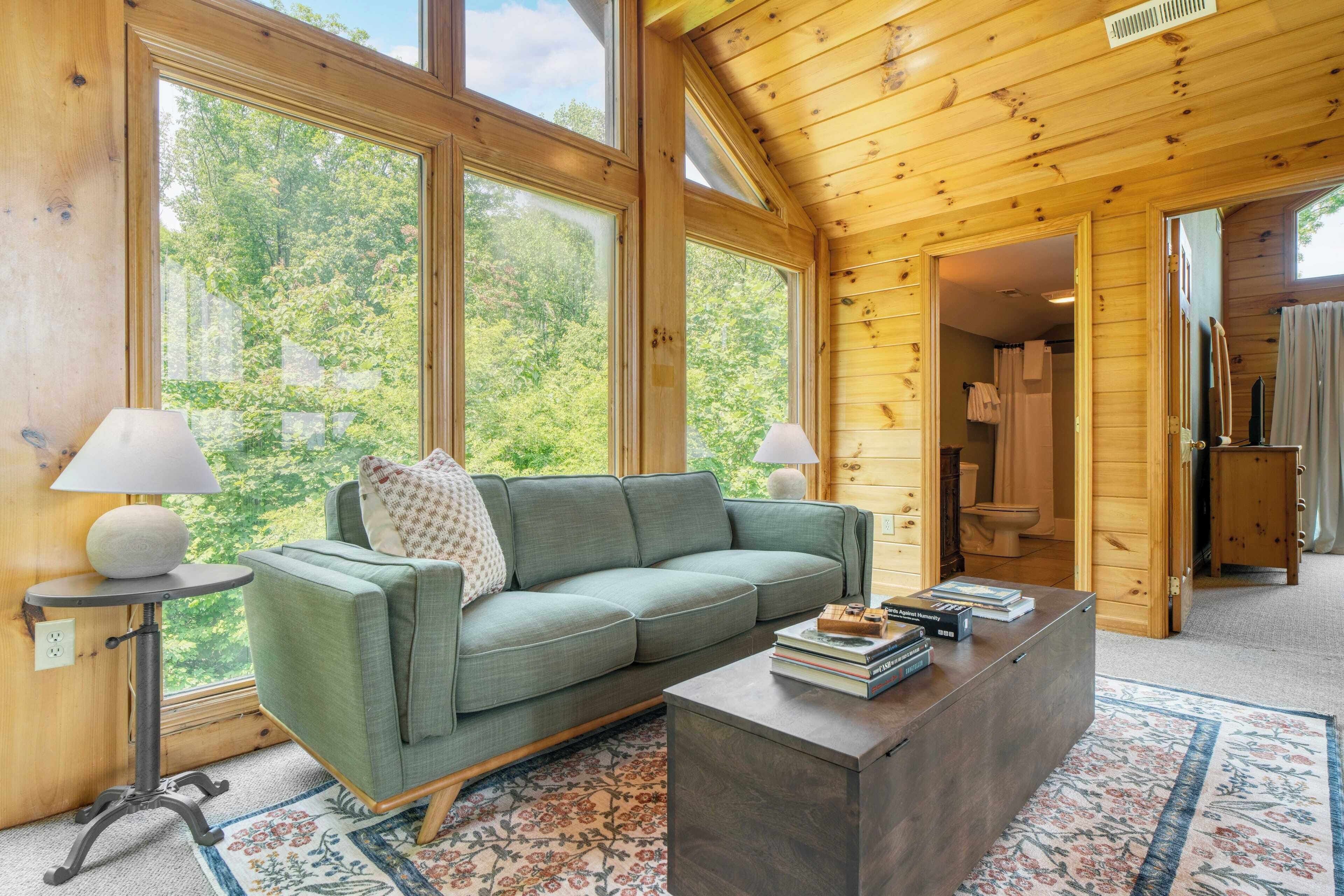 A cozy cabin interior with a green sofa, a coffee table with books, and large windows offering a view of the forest.