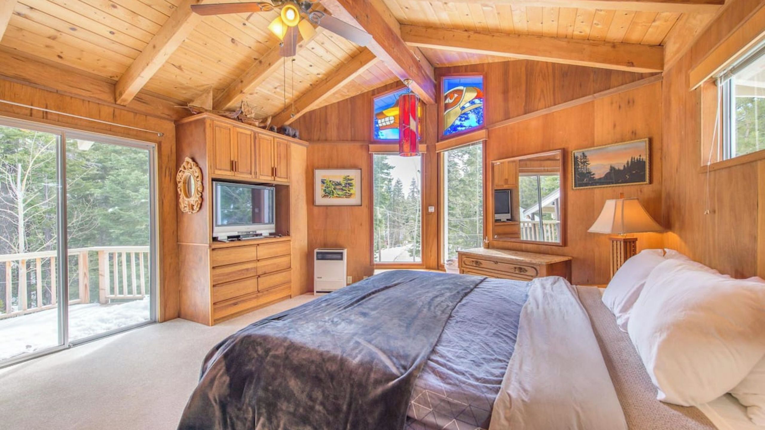 A wooden cabin bedroom with a bed, dresser, TV, and large windows.