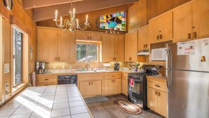 A kitchen with wooden cabinets, a stainless steel refrigerator, a microwave, a stove, and a chandelier. - Mahogany by Avantstay Snowbird Cabin w/ Full Chamberlands Access! (Homewood)