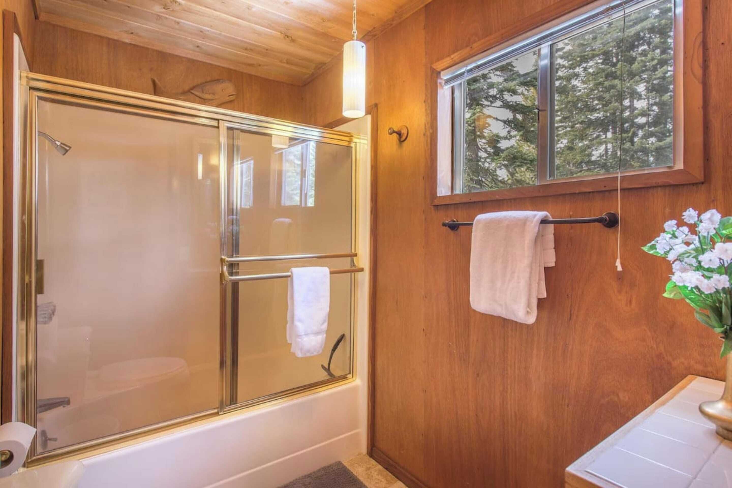 A wooden interior with a glass-enclosed shower, a window with a view of trees, and a towel rack with towels.
