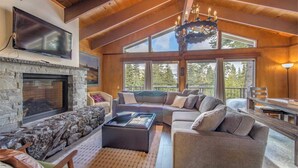 A spacious living room with a stone fireplace, a large flat-screen TV, a cozy sofa, and a dining area with a wooden table. - Mahogany by Avantstay Snowbird Cabin w/ Full Chamberlands Access! (Homewood)
