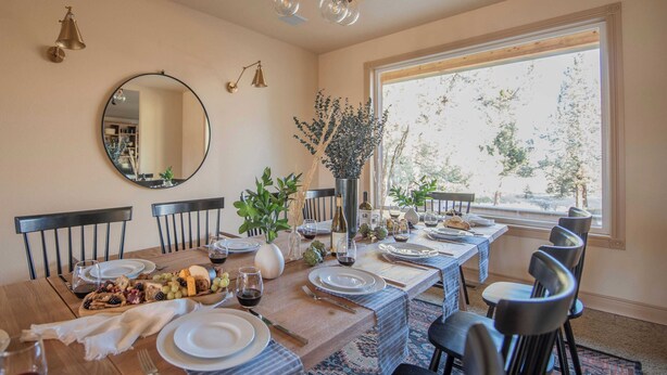 Une salle à manger avec une longue table dressée pour un repas, entourée de chaises, et une grande fenêtre donnant sur la forêt.