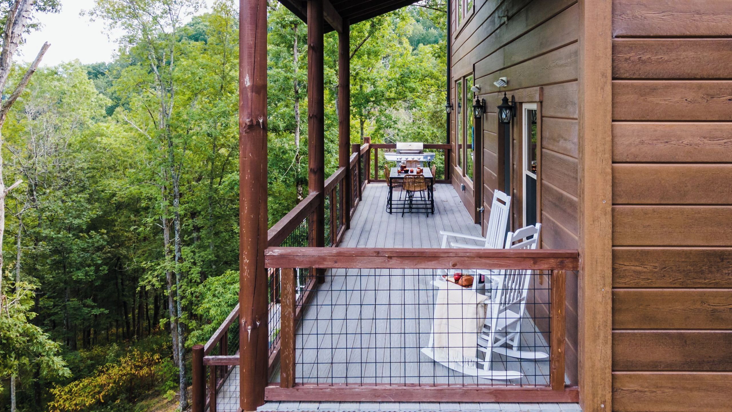 A wooden deck with a table and chairs, surrounded by a wooden railing, overlooking a forested area.