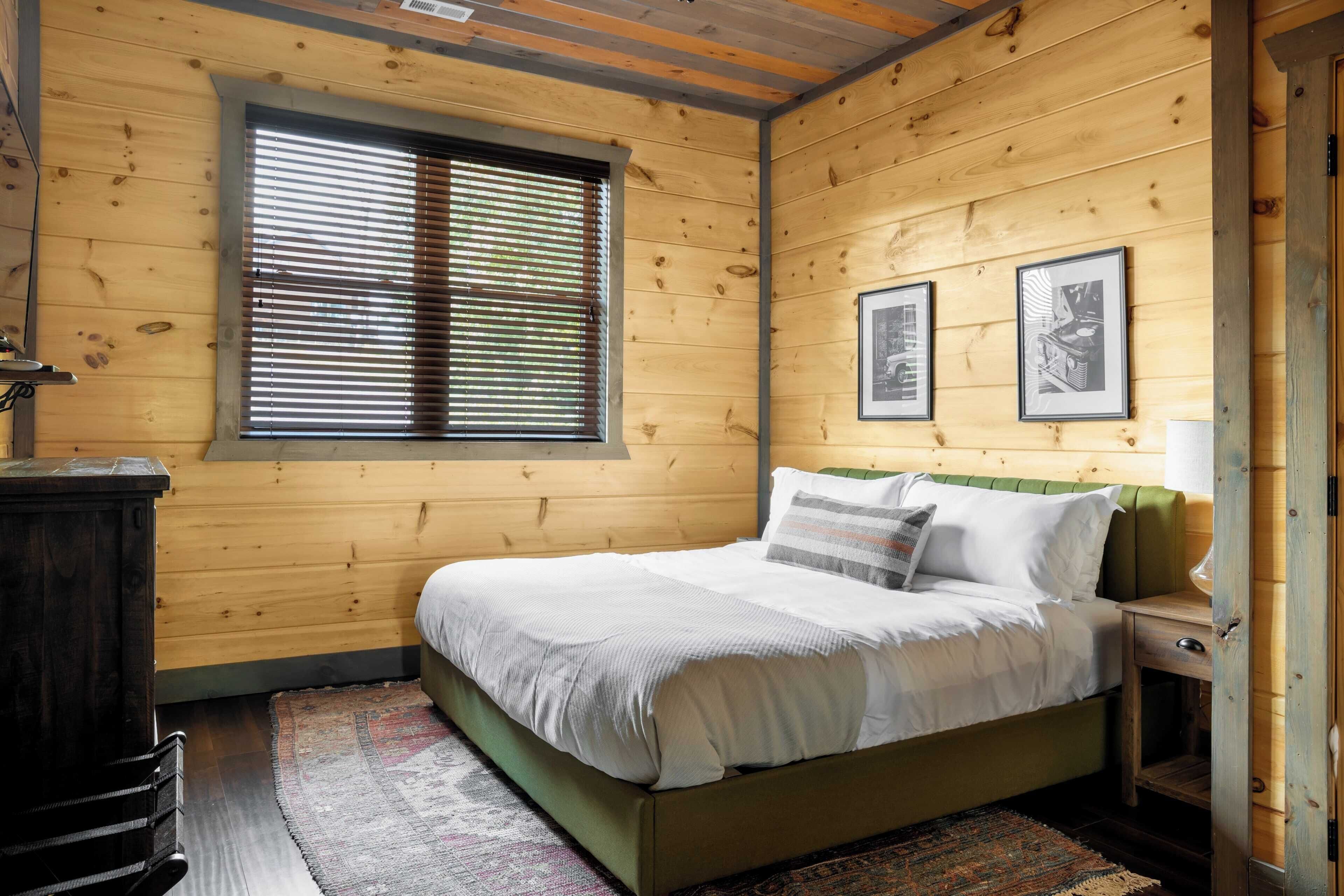 Une chambre aux murs lambrés, avec un lit, une fenêtre équipée de stores et deux tableaux encadrés accrochés au mur.