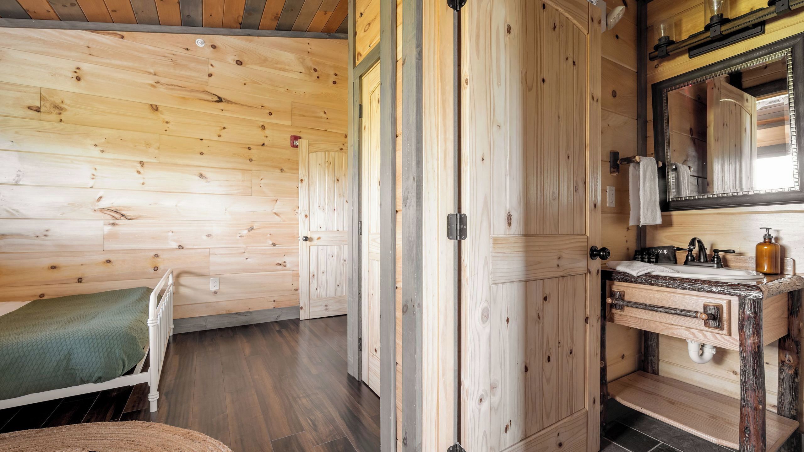 A wooden cabin interior with a bed, a wooden wardrobe, a bathroom with a sink and mirror, and a window.
