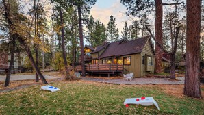 A lodge with a deck and outdoor seating area surrounded by tall trees. - Walk to the Lake, Sand Volleyball Court, Pool Table, Deck Gambler by Avantstay (Big Bear Lake)