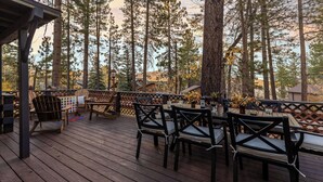 A wooden deck with outdoor seating, surrounded by tall trees. - Walk to the Lake, Sand Volleyball Court, Pool Table, Deck Gambler by Avantstay (Big Bear Lake)