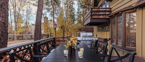 Un área de comedor al aire libre con una parrilla, terraza de madera y árboles circundantes.