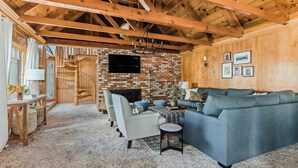 A spacious living room with a brick fireplace, wooden beams, a large sofa, and a television mounted on the wall. - Walk to the Lake, Sand Volleyball Court, Pool Table, Deck Gambler by Avantstay (Big Bear Lake)