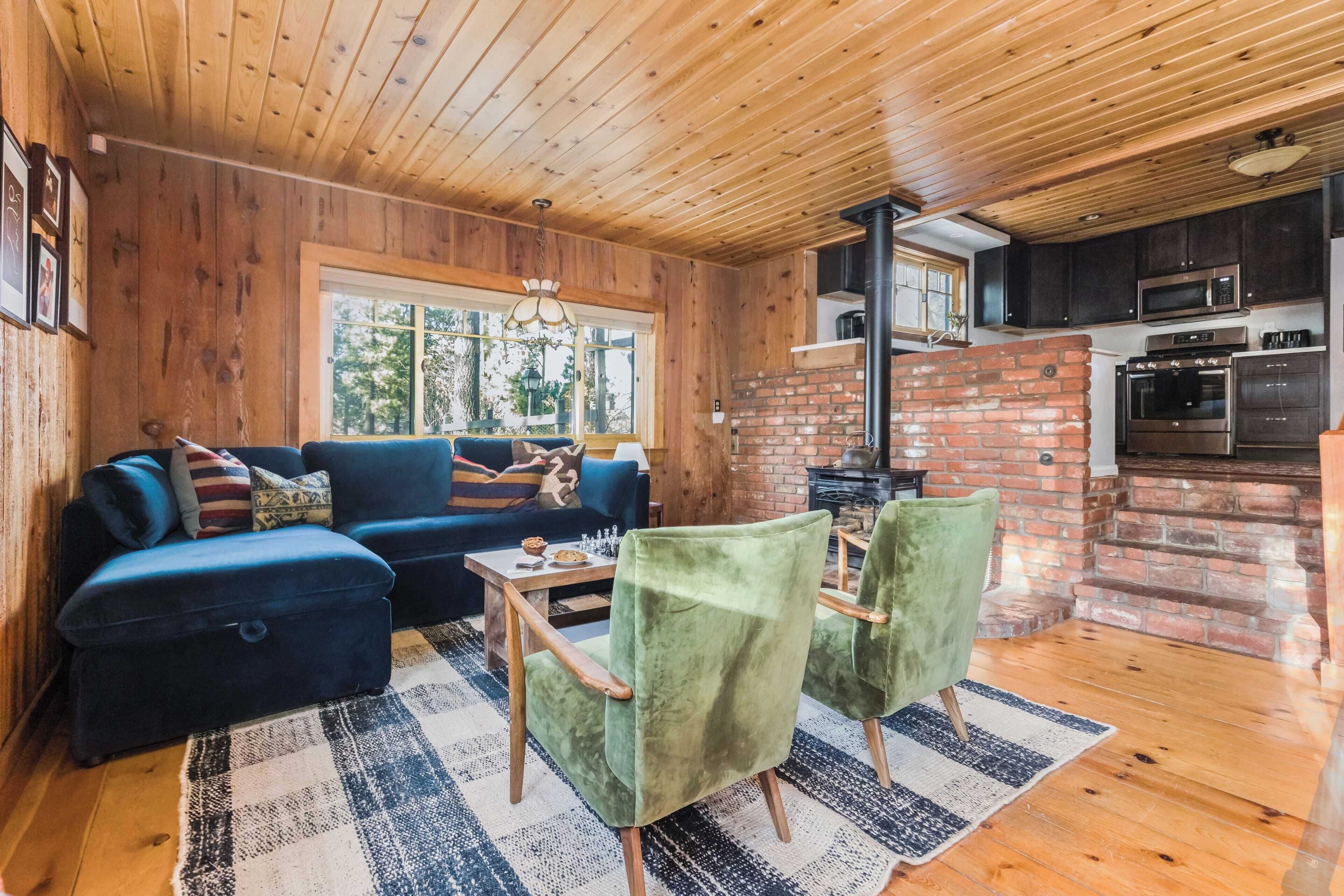Ein gemütliches Blockhaus-Interieur mit Holzbalkendecke, Kamin aus Ziegelsteinen, einer Couch, zwei grünen Sesseln und einer Küchenzeile.
