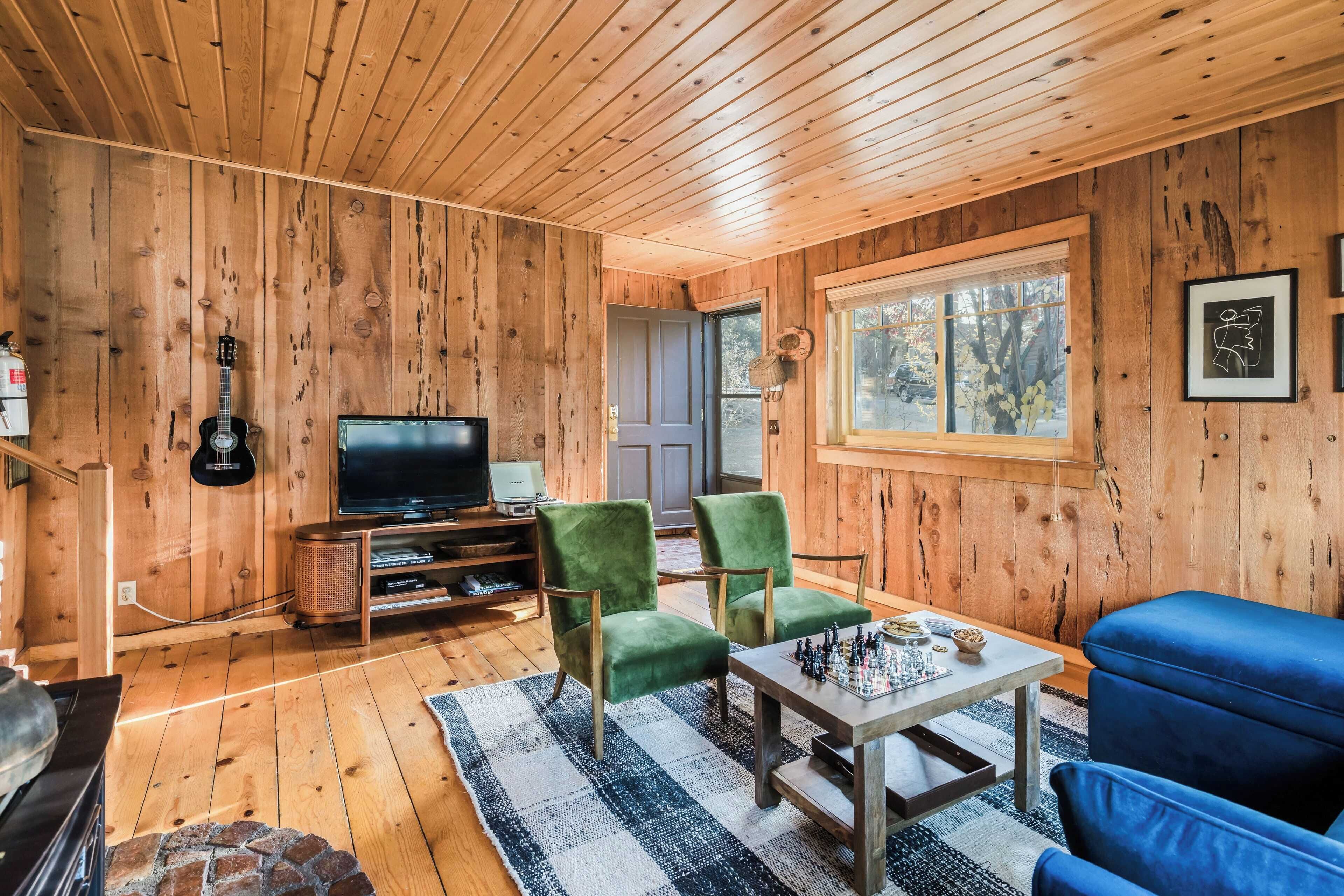 A wooden-paneled room with a television, green and blue chairs, a chess table, and a stone fireplace.