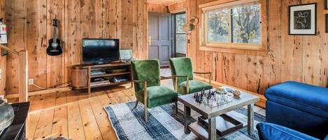 A wooden-paneled room with a television, green and blue chairs, a chess table, and a stone fireplace.