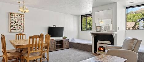 A living room with a wooden dining table, chairs, a flat-screen TV, a fireplace, and a large window with a mountain view.