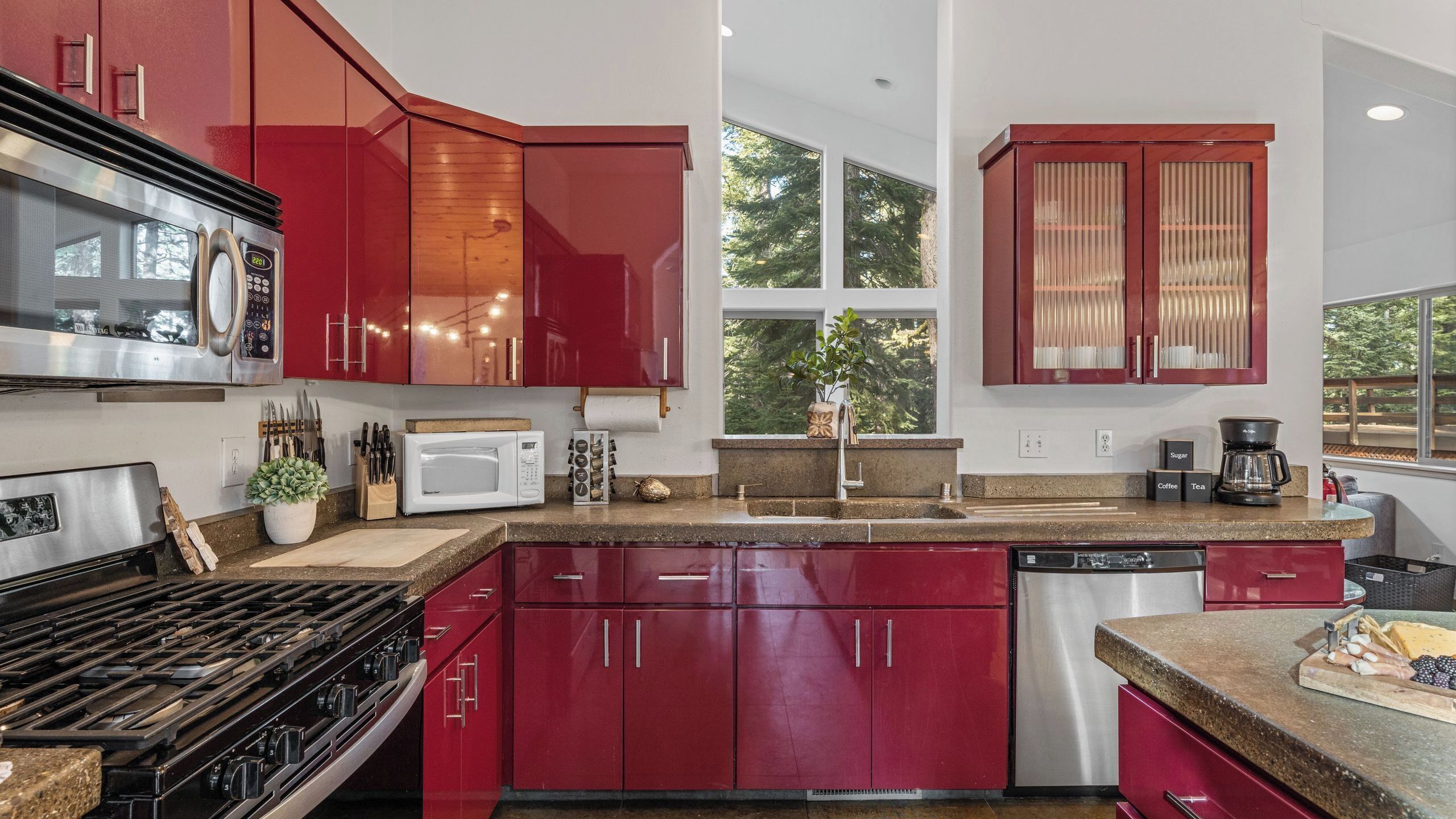 A modern kitchen with red cabinets, stainless steel appliances, and a large window providing natural light.