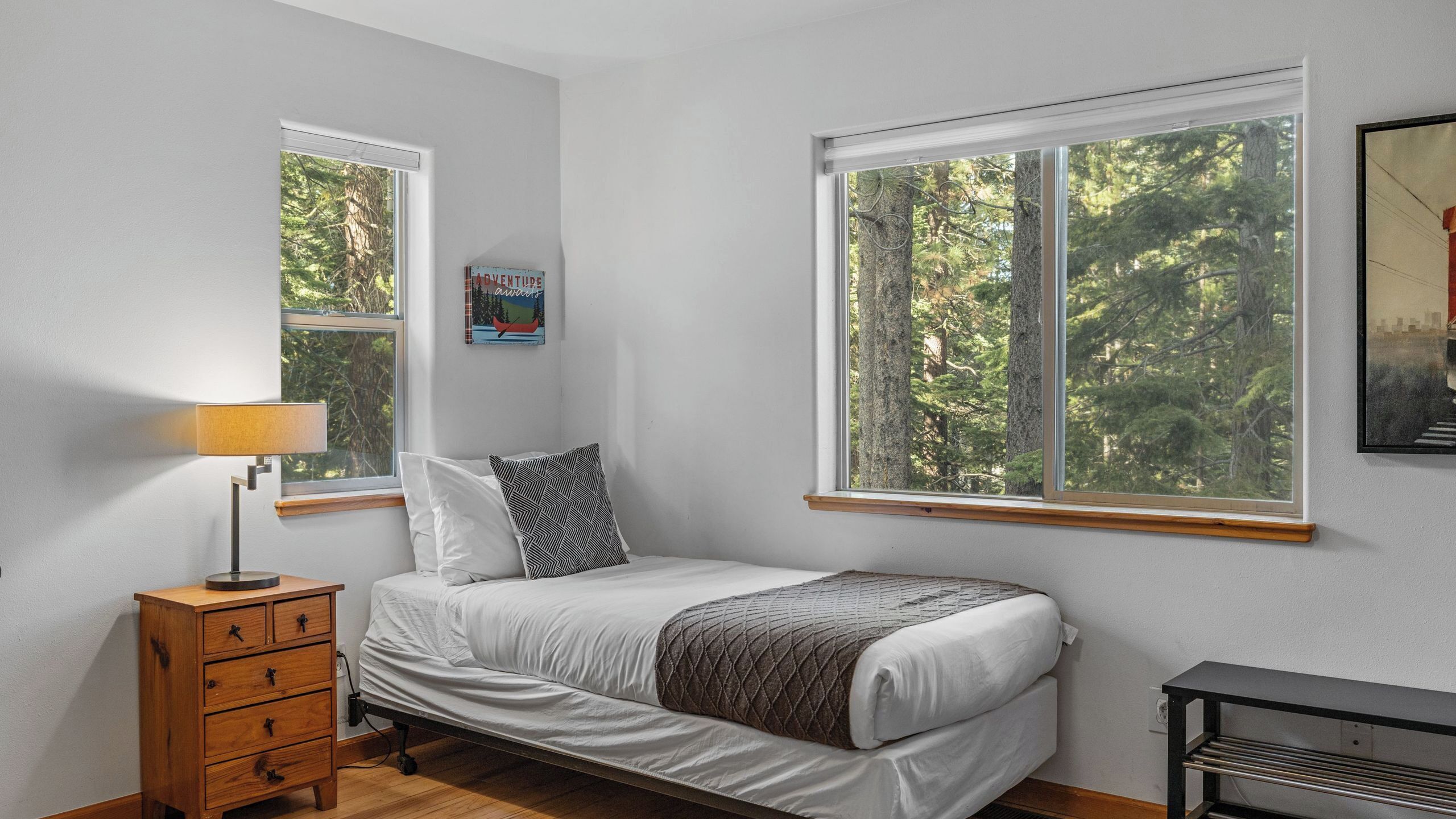 Une chambre à coucher comprenant un lit, une table de chevet, une lampe, une commode et deux fenêtres donnant sur une forêt.