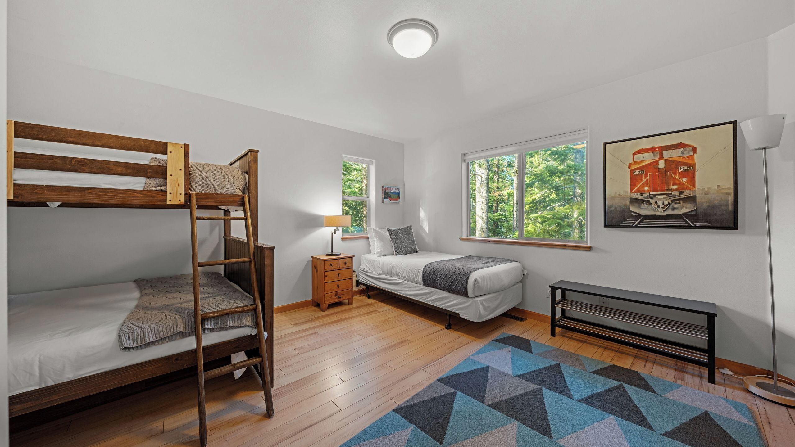 Une chambre avec un lit superposé, un lit simple, une table de chevet, une lampe, un radiateur, une fenêtre donnant sur des arbres et un tableau encadré au mur.